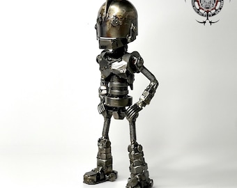 May include: A metallic robot sculpture constructed from various industrial hardware components including nuts, bolts, and springs. The figure features a rounded head with a crest, a torso with visible joints, and thin legs, all finished in a dark, polished steel.