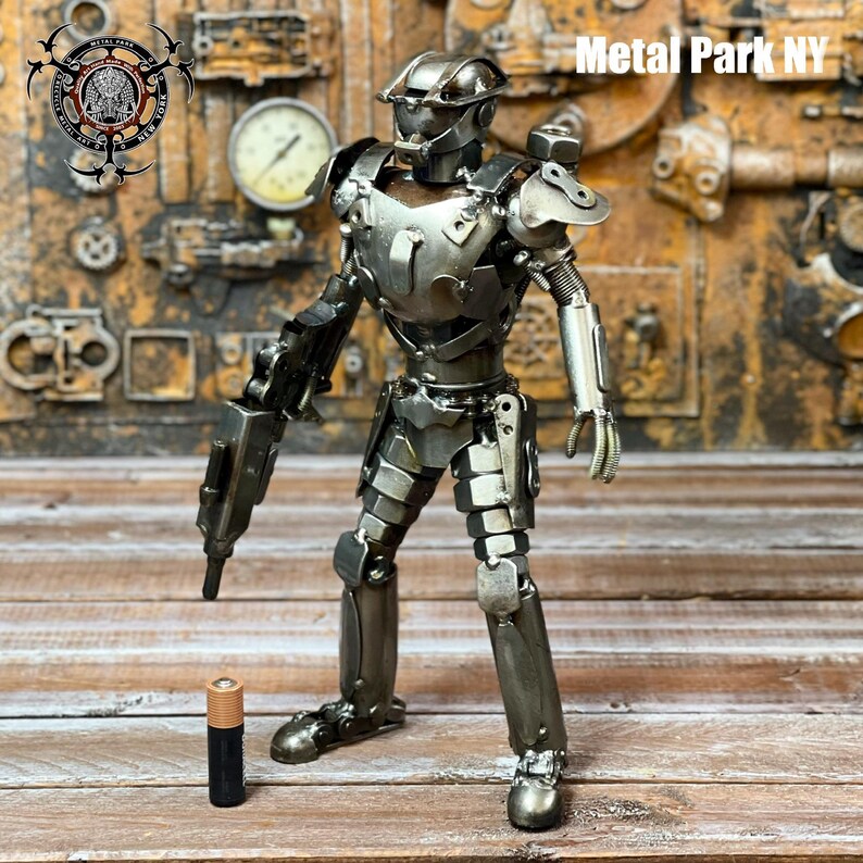 May include: A handcrafted metal robot sculpture with a gun, made from various metal pieces. The sculpture is silver and gray, with visible welds and textures. A standard AA battery is placed next to the robot for scale. The text "Metal Park NY" is visible.