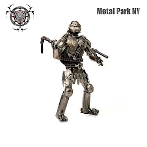 May include: A metal sculpture of a figure with a helmet, chain belt, and two guns. The sculpture is made of various metal pieces welded together. The figure is standing on a white background. The text "Metal Park NY" is above the figure.