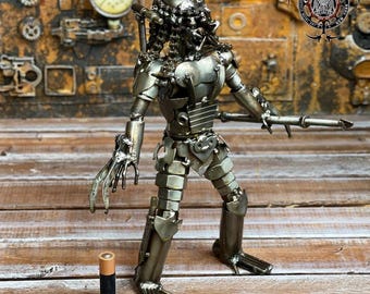 May include: A metallic sculpture of a Predator figure, crafted from various metal pieces, including nuts and bolts. The figure is posed with a weapon and has intricate details. A battery is placed near the figure for scale. The background features industrial-themed elements.