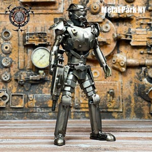 May include: A handcrafted metal sculpture of a futuristic soldier. The figure is made from various metal pieces, including nuts, bolts, and plates, and is holding a weapon. The background features industrial elements, and the text "Metal Park NY" is visible.