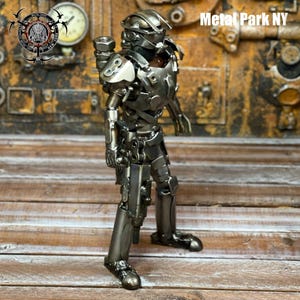 May include: A metallic sculpture of a futuristic soldier, crafted from various metal components. The figure is posed in a standing position, showcasing intricate details and a polished finish. The sculpture is set against a backdrop with the text "Metal Park NY".