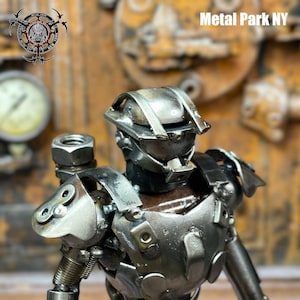 May include: A detailed metal sculpture of a humanoid figure, constructed from various metal components. The sculpture has a helmet, body armor, and articulated limbs. The text "Metal Park NY" is visible in the background.