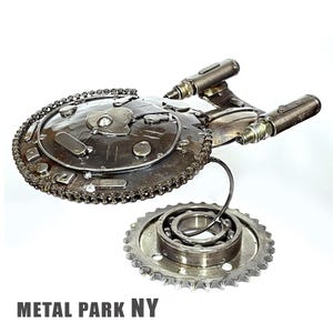 May include: A metal sculpture of the USS Enterprise starship from Star Trek. The sculpture is made from recycled metal parts, including gears, chains, and bolts. The sculpture is painted in a silver color and has a weathered finish. The sculpture is signed "METAL PARK NY".