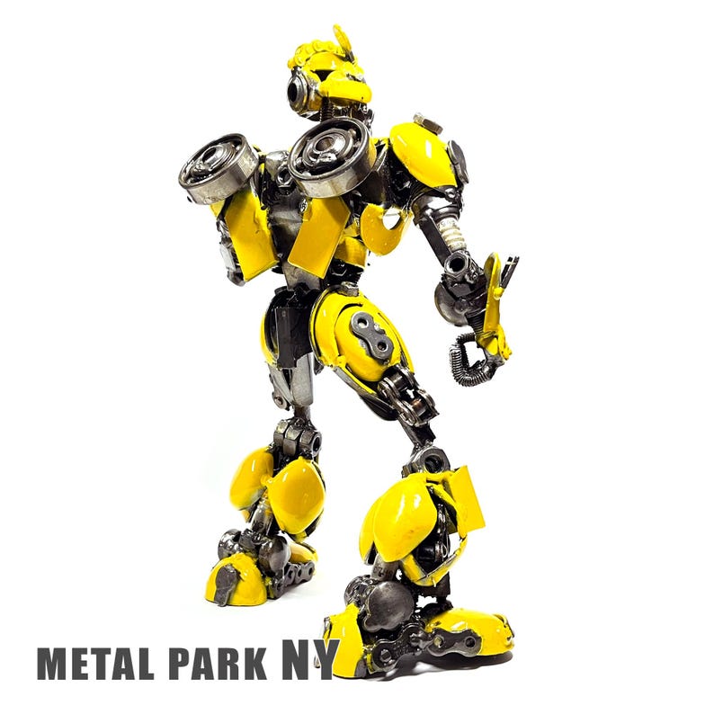 May include: A robot sculpture constructed from recycled metal parts, featuring a bright yellow finish on its armour plates and exposed mechanical joints. The figure stands in a dynamic pose with visible gears and bolts. Text reads METAL PARK NY.