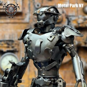 May include: A handcrafted metal sculpture of a humanoid robot. The robot is constructed from various metal pieces, including plates, bolts, and springs, with a silver finish. The sculpture has the text "Metal Park NY".