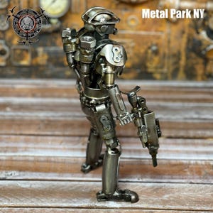 May include: A handcrafted metal sculpture of a robot soldier. The figure is constructed from various metal parts, including nuts, bolts, and other hardware. The sculpture is silver and has a gun in its right hand. The text "Metal Park NY" is visible.