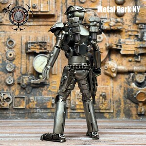 May include: A handcrafted metal sculpture of a humanoid figure, constructed from various metal components. The sculpture has a silver finish and is posed in a standing position. The background features a textured, rusty metal wall. The text "Metal Park NY" is visible.