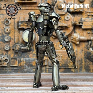 May include: A handcrafted metal robot sculpture. The robot is constructed from various metal parts, including bolts, nuts, and gears. It has a detailed design with a helmet, shoulder pads, and a weapon. The sculpture is a unique piece of art with the text "Metal Park NY".