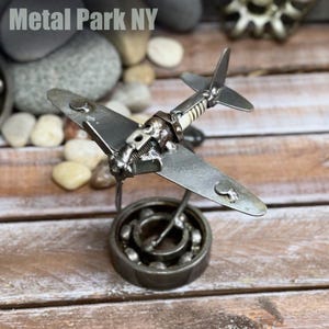 May include: A handcrafted metal airplane sculpture. The wings and body are made of silver-colored metal, with a propeller and tail. The sculpture is mounted on a bearing base. The text "Metal Park NY" is in the top left corner.