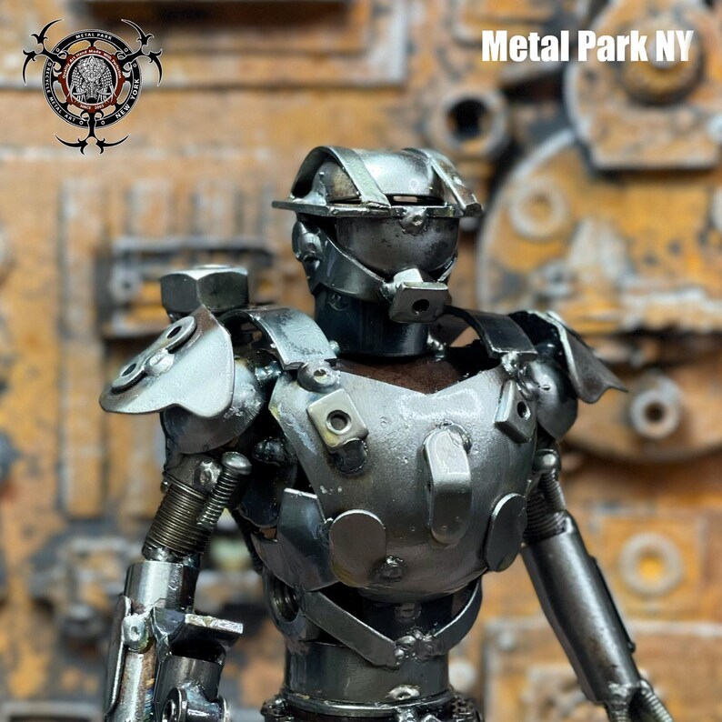 May include: A handcrafted metal sculpture of a humanoid figure, constructed from various metal pieces. The figure has a helmet, chest armor, and articulated arms. The text "Metal Park NY" is visible. The sculpture is set against a textured background.