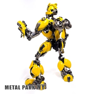 May include: A robot figurine constructed from recycled metal parts, featuring a bright yellow and metallic grey colour scheme. The figure has a humanoid form with articulated joints, circular gear components on the chest, and the text METAL PARK NY.