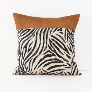 Puede incluir: Una almohada cuadrada con tejido de estampado de cebra en blanco y negro. La almohada tiene un ribete de imitación de cuero marrón alrededor del borde superior.