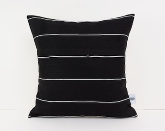 Könnte beinhalten: Quadratisches schwarzes Kissen mit horizontalen weißen Streifen. Das Kissen hat ein schlichtes, modernes Design und ist aus einem strukturierten Stoff gefertigt. Ein dekorativer Artikel für ein Bett oder Sofa.