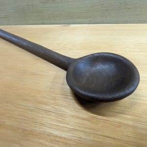 Könnte beinhalten: Ein dunkelbrauner Holzlöffel mit langem Stiel und rundem Löffel. Der Löffel besteht aus einem einzigen Holzstück und hat eine glatte, polierte Oberfläche. Der Löffel liegt auf einer hellbraunen Holzoberfläche.