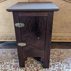 May include: A dark brown wooden nightstand with a single door and brass hinges. The nightstand has a simple design with a slightly curved base.