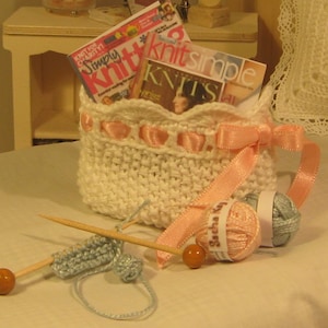 May include: A miniature white crocheted basket with pink ribbon accents and a bow, holding two magazines labeled "Simply Knit" and "Knits Simple." Knitting needles, yarn, and a small knitted swatch are nearby, suggesting a crafting theme.