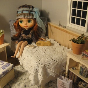 May include: A doll wearing a blue and brown knitted hat and a black dress sits on a white crocheted bedspread. The doll has blue eyes and long red hair. The doll is in a dollhouse bedroom with a white nightstand, a potted plant, and a bookshelf.