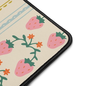 May include: A corner view of a beige mousepad with a black stitched edge. The mousepad features a repeating pattern of pink strawberries with green tops, orange flowers, and green vines. A light blue and yellow decorative border is visible.