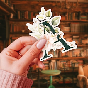 Puede incluir: Una pegatina con la letra "A" en color verde oscuro, adornada con elementos florales y botánicos. El diseño incluye flores de color rosa claro, hojas verdes y delicados detalles dorados, creando una estética decorativa y artística.
