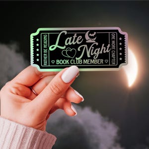 Pode incluir: Um autocolante preto e iridescente em forma de bilhete, segurado por uma mão. O autocolante diz "Late Night Book Club Member" com um gráfico de lua e morcego. O autocolante tem uma borda verde e o texto "Rather be reading" e "One more chapter".
