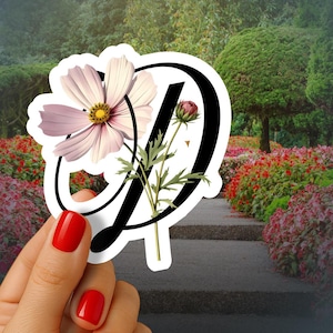 May include: A sticker featuring the letter "D" in black, adorned with a light pink and white flower and a bud. The sticker has a white border. The background shows a garden with a stone pathway and red flowers.