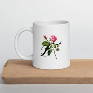 Può includere: Tazza in ceramica bianca con una rosa rosa e la lettera "A" in nero. Il design floreale comprende foglie verdi e un bocciolo di rosa. La tazza è su una superficie di legno, ideale per caffè o tè.