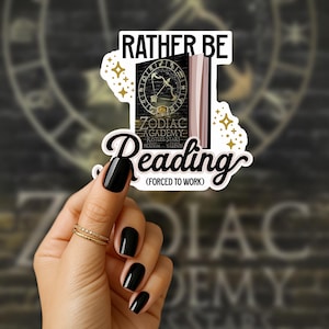Peut inclure: Un autocollant avec le texte "RATHER BE READING (FORCED TO WORK)". L'autocollant présente un livre avec le titre "ZODIAC ACADEMY" et un motif doré de flèche et arc. L'autocollant est tenu par une main avec du vernis à ongles noir.