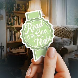 May include: A light green sticker with the text "Read It In One Night" in a whimsical font. The sticker is held in front of a cozy room with a bookshelf and armchair.