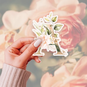 May include: A sticker in the shape of the letter "A", decorated with a floral design. The sticker features blush pink flowers, green leaves, and gold glitter. The background is a blurred image of pink roses.