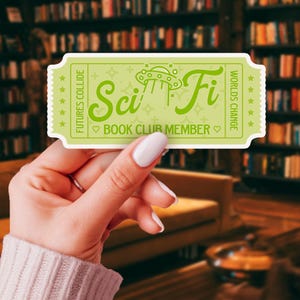 Pode incluir: Um autocolante verde claro com um design de bilhete, com as palavras "Sci Fi" numa fonte estilizada, juntamente com uma ilustração de OVNI. O texto "Book Club Member" também está incluído. O autocolante tem uma borda serrilhada e as palavras "Futures Collide" e "Worlds Change".