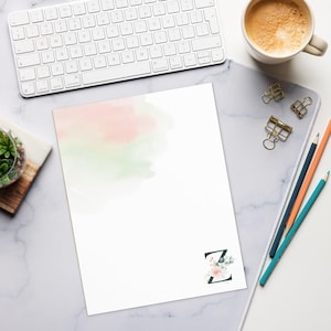 Può includere: Carta bianca con un lavaggio ad acquerello rosa e verde. Un bordo dorato incornicia la parte superiore della carta. La lettera "Z" è decorata con fiori e verde. Sono visibili anche una tastiera, matite e una tazza di caffè.