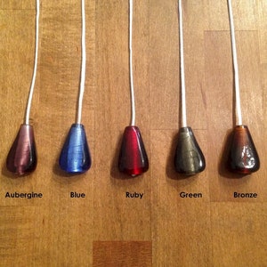 May include: Five glass light pull chains in different colours: aubergine, blue, ruby, green, and bronze. The chains are hanging from a white cord.