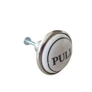 May include: A white ceramic cabinet knob with the word "PULL" in black lettering. The knob has a silver metal rim and a silver screw for installation. This decorative hardware is suitable for drawers and cabinets.