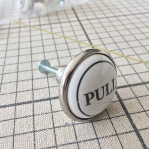 Könnte beinhalten: Weißer Keramik-Schrankknopf mit silbernem Metallrand. Der Knopf hat einen schwarzen Kreis mit dem Wort "PULL" in Schwarz.