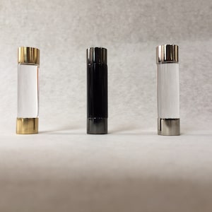 May include: Three decorative bottle stoppers, one with a gold top, one with a black top, and one with a silver top. Each stopper is made of clear glass and has a cylindrical shape.