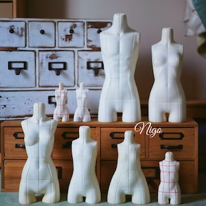 May include: A collection of white dress forms of various sizes, arranged on a wooden surface. Some forms are solid white, while others have a red grid pattern. The forms are displayed in front of a vintage wooden cabinet.