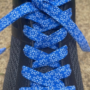 May include: Close-up of a black athletic shoe with bright blue and white patterned shoelaces. The laces feature a repeating floral design. The shoe is laced up and ready to wear.