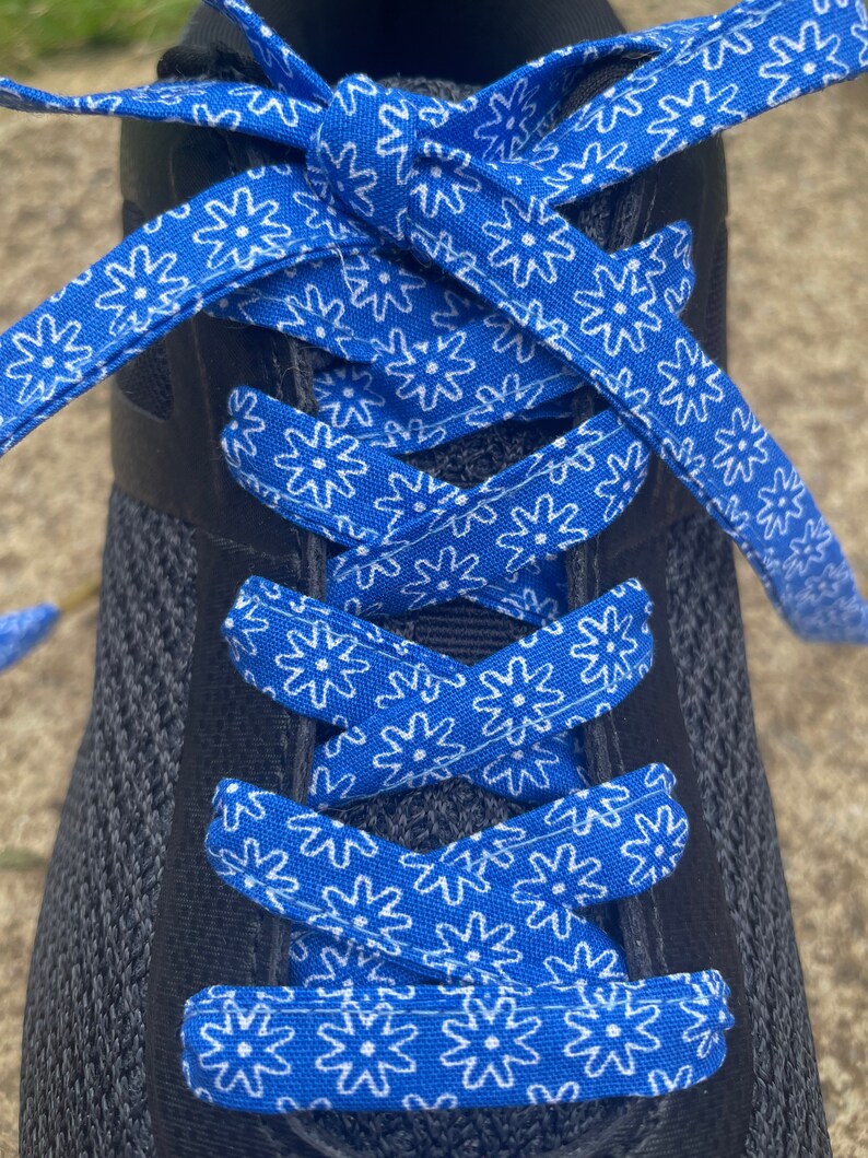 May include: Close-up of a black athletic shoe with bright blue and white patterned shoelaces. The laces feature a repeating floral design. The shoe is laced up and ready to wear.