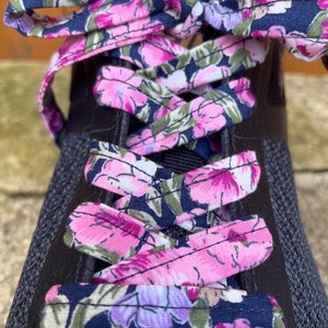 Könnte beinhalten: Nahaufnahme eines schwarzen Sportschuhs mit floralen Schnürsenkeln. Die Schnürsenkel haben einen marineblauen Hintergrund mit rosa, lila und grünen Blumenmustern. Die Schnürsenkel sind zu einer Schleife gebunden.