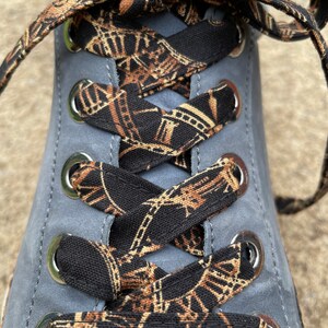 May include: A grey sneaker featuring flat black shoelaces with a repeating gold-toned clock face and Roman numeral pattern. The laces are threaded through polished silver-colored metal eyelets, creating a crisscross design across the shoe tongue.