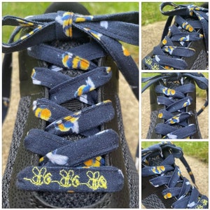 Puede incluir: Primer plano de una zapatilla deportiva negra con cordones azul marino. Los cordones tienen un estampado de flores amarillas, blancas y azules. La lengüeta del zapato tiene tres abejas amarillas bordadas.