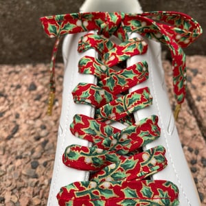 Puede incluir: Una zapatilla blanca con cordones de tela planos que presentan un patrón festivo de hojas de acebo y bayas en rojo, verde y blanco. Los cordones están atados en un lazo en la parte superior y tienen herretes metálicos dorados.