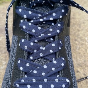 以下が含まれることがあります： ネイビーブルーのドット柄の靴紐が付いた黒いスニーカーのクローズアップ。靴紐はクロス模様に結ばれており、ネイビーブルーの背景に白い水玉模様があります。靴は黒いメッシュアッパーです。