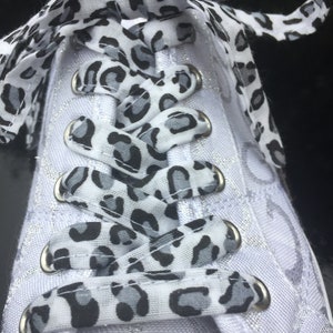 May include: Close-up of a white sneaker with silver accents and leopard print laces. The laces are black, white, and gray. The eyelets are silver. The shoe is a low-top style.