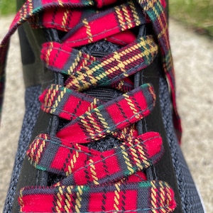 May include: A black athletic shoe featuring flat, wide shoelaces with a red, green, and gold plaid pattern. The laces are threaded through the eyelets in a crisscross design, contrasting against the dark, textured mesh and synthetic upper material.