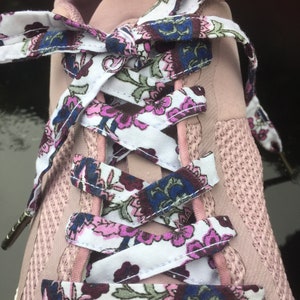 Pode incluir: Close-up de um tênis rosa com cadarços estampados florais. Os cadarços apresentam um fundo branco com um padrão repetido de flores rosa, azuis e bordô. O tênis tem uma textura de malha.
