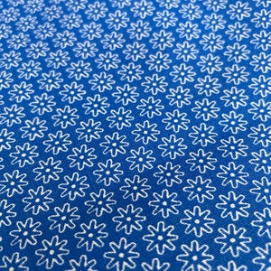 May include: Close-up of a blue fabric with a repeating white floral pattern. The flowers have a simple, stylized design, creating a textured appearance. The fabric's color is a vibrant, solid blue.