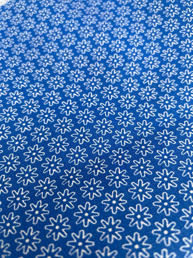 May include: Close-up of a blue fabric with a repeating white floral pattern. The flowers have a simple, stylized design, creating a textured appearance. The fabric's color is a vibrant, solid blue.