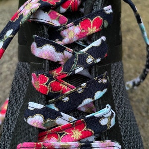 May include: Close-up of a black trainer with floral patterned shoelaces. The laces feature a repeating pattern of pink, red, and gold flowers on a navy blue background. The laces are threaded through the eyelets of the shoe.